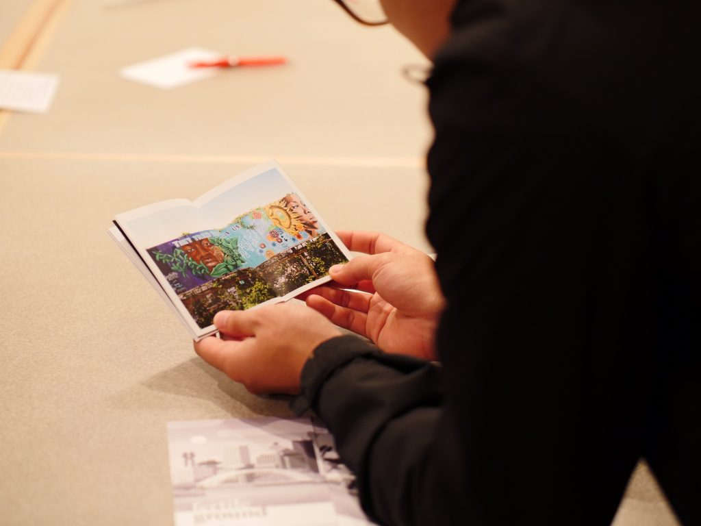 a gallery visitor leans on a table and holds volume one of the fertile ground zine open to a spread featuring a photograph of a mural