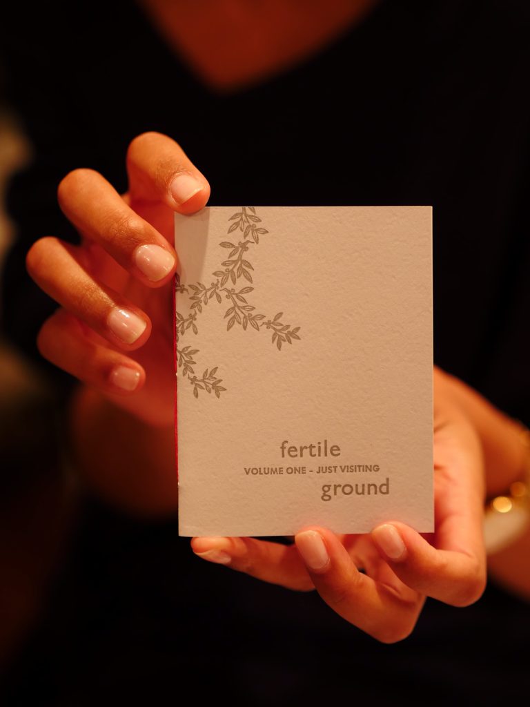 kate holds a copy of volume one of the fertile ground zine: a passport-sized light gray book with leaves and text on the cover, bound with red thread